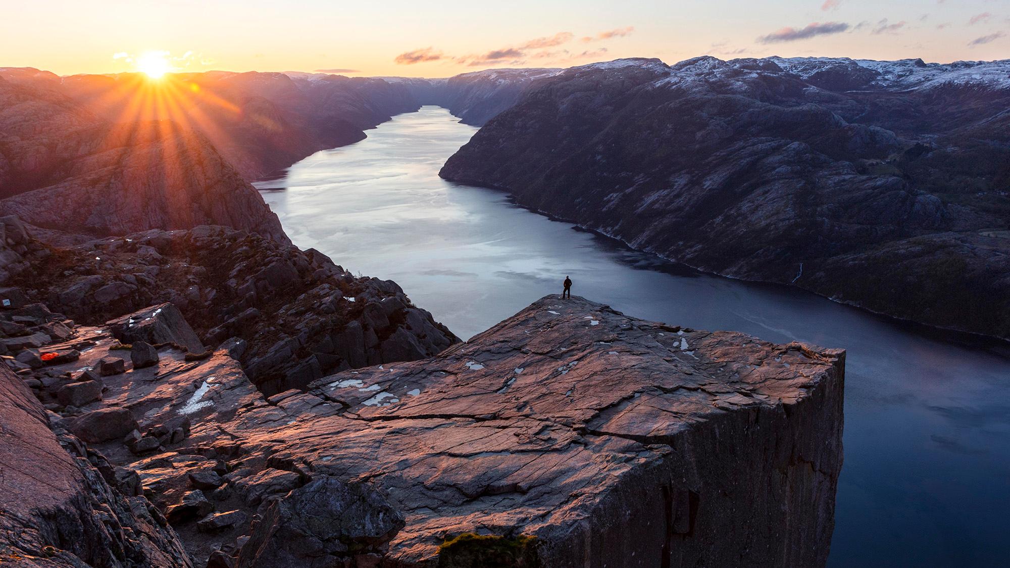 En person på kanten av Preikestolen mens sola skimtes bak fjellene.