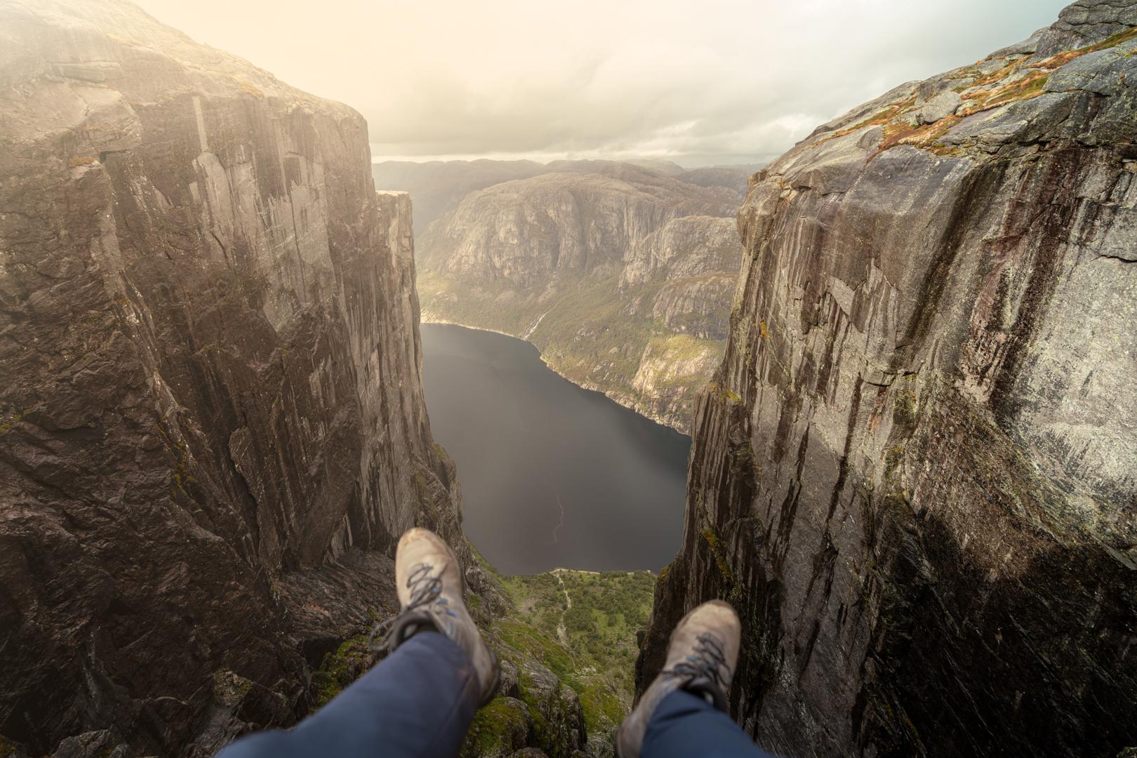 HIker's feet dangling from a viewing point of the Lysefjord. Scenic views with massive mountains on either side and the fjord in the middle.